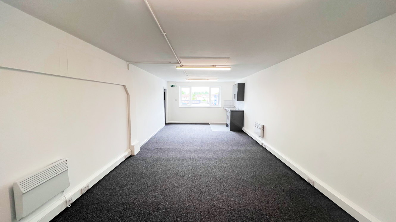 Empty rectangular room with white walls, grey carpet, a window at the far end, overhead fluorescent lights, and a small kitchenette in the back right corner.