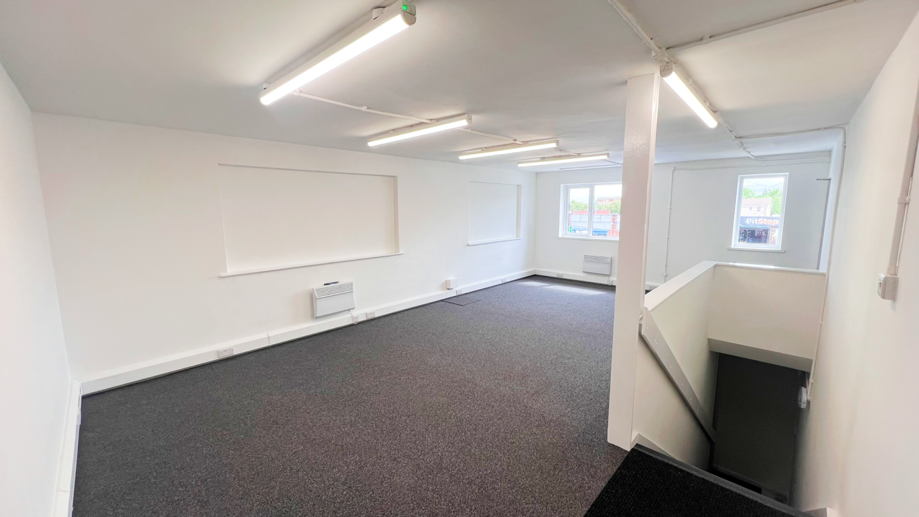 A clean, empty office space with white walls, grey carpet, fluorescent lighting, two windows, and a staircase leading to a lower floor.
