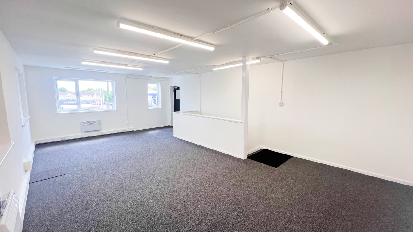 Bright, empty office space with white walls, grey carpet, fluorescent ceiling lights, two windows, and a partial room divider near the centre.