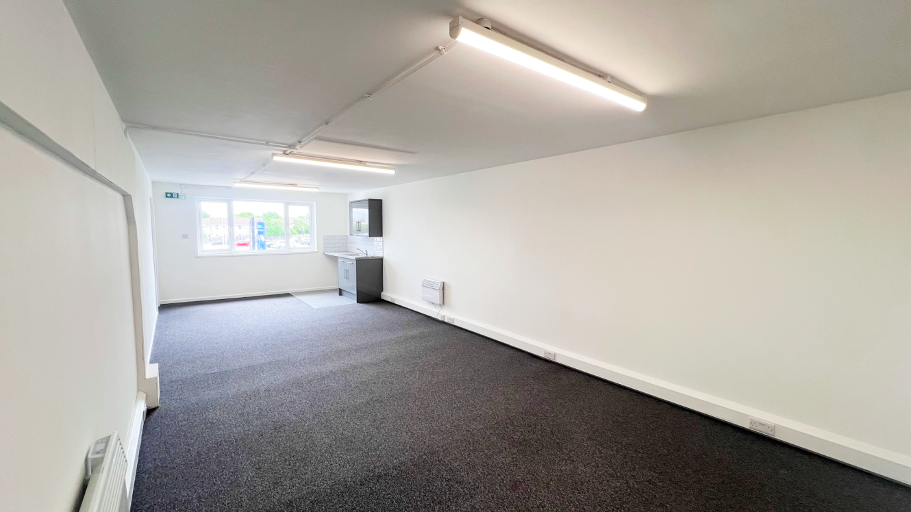 A bright, empty office space with white walls, grey carpet, fluorescent lights, a small kitchenette, and a window at the far end.