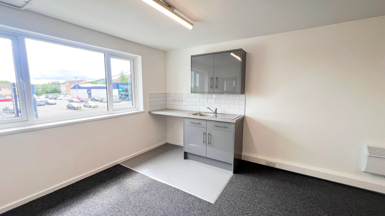 Small kitchenette with grey cupboards, white tiled splashback, and a sink in a bright room that has a large window looking out over a car park.