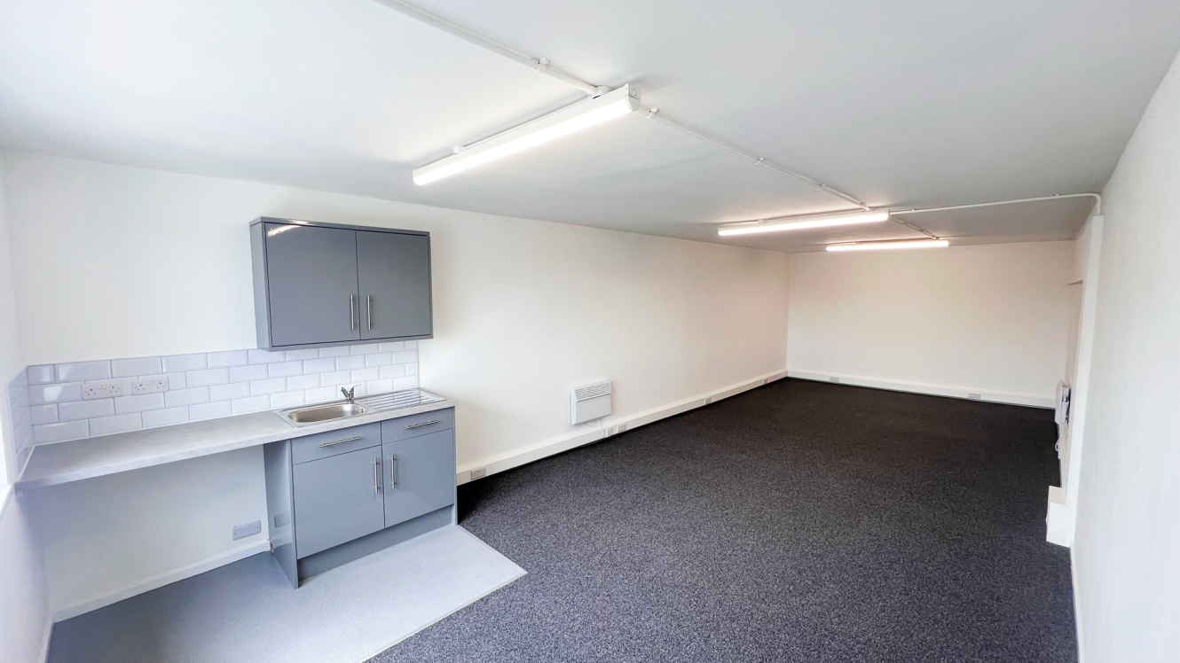 A small, empty office with white walls, dark carpet, overhead fluorescent lights, and a grey kitchenette with a sink and cupboards on the left side.