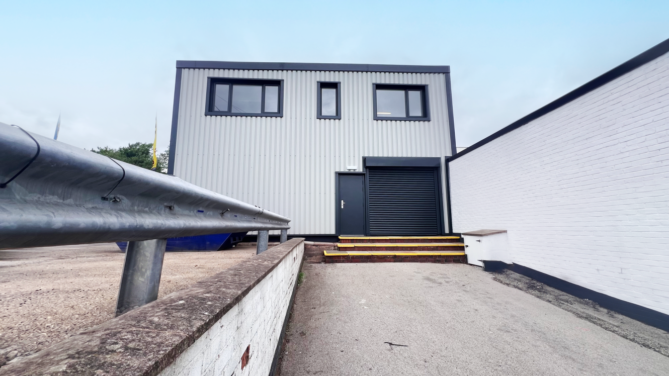 A two-storey industrial building with grey metal cladding and black window frames stands next to a white brick wall, featuring a metal railing along a concrete drive.