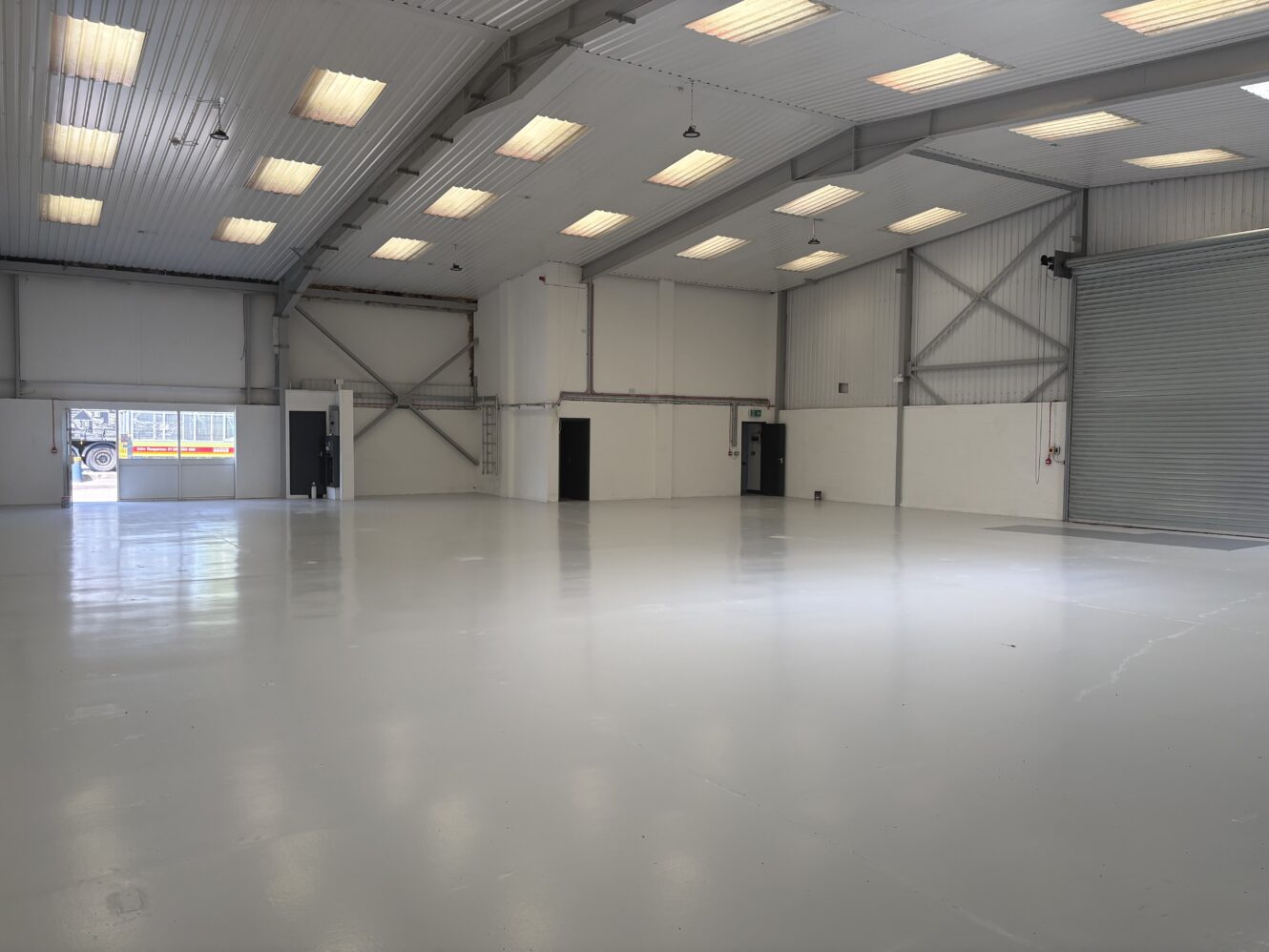 Large empty warehouse with bright overhead lights, white walls, high ceilings, and two large doors, one open and one closed.