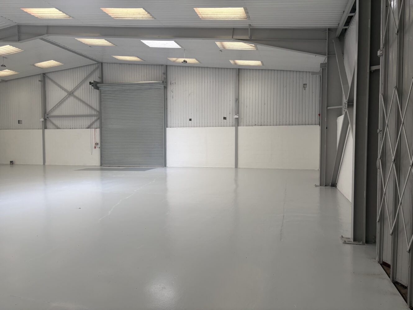 Empty industrial warehouse with a polished concrete floor, metal walls, overhead lights, and a large closed roller shutter door.