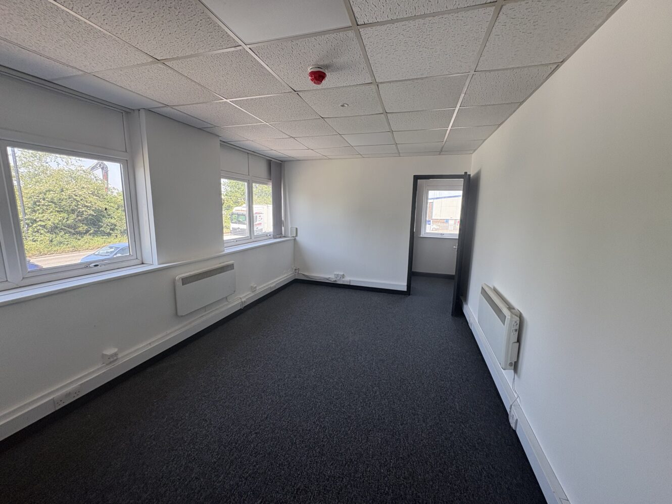 Empty office room with white walls, three windows on the left side, dark carpeted floor, ceiling tiles, wall heaters, and an open doorway at the far end.