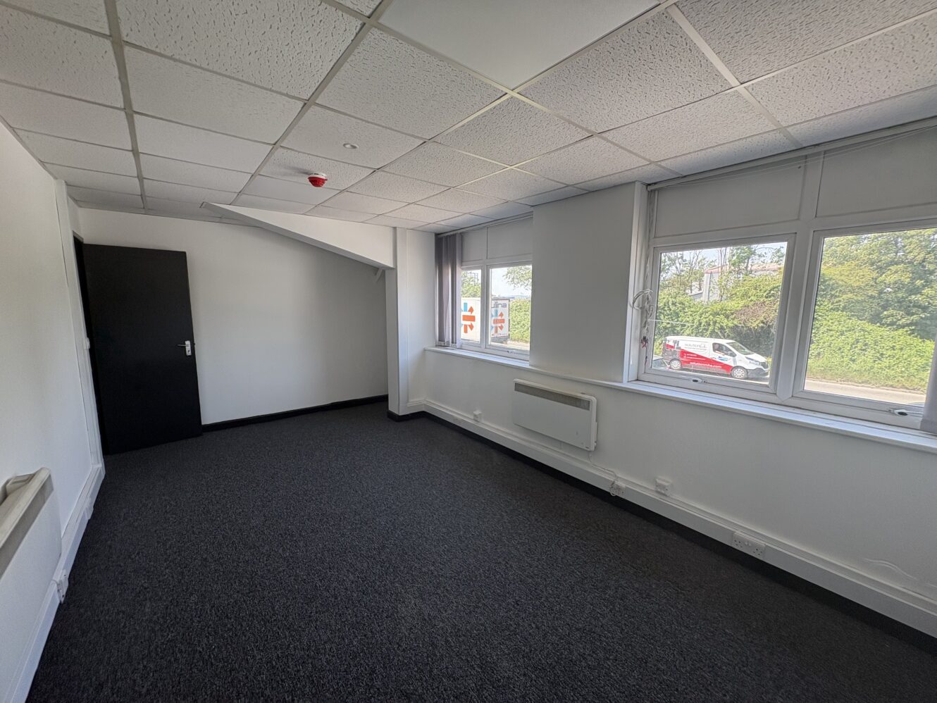 Empty office room with grey carpet, white walls, black door, wall heater, and large windows overlooking a street with a red car and greenery outside.