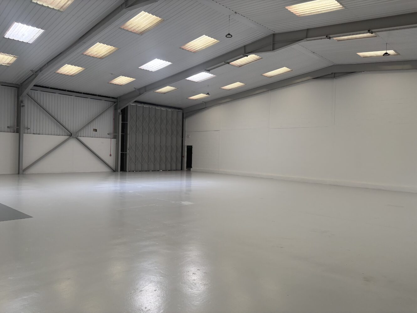 Empty industrial warehouse with white walls, high ceilings, and overhead lighting; metal beams and a large folding door are visible on one side.