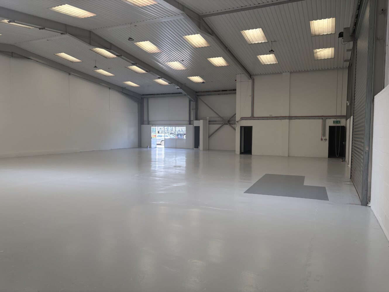 Large empty warehouse with white walls, high ceiling, bright fluorescent lighting, and a polished floor. Several doors and an open shutter are visible at the far end.