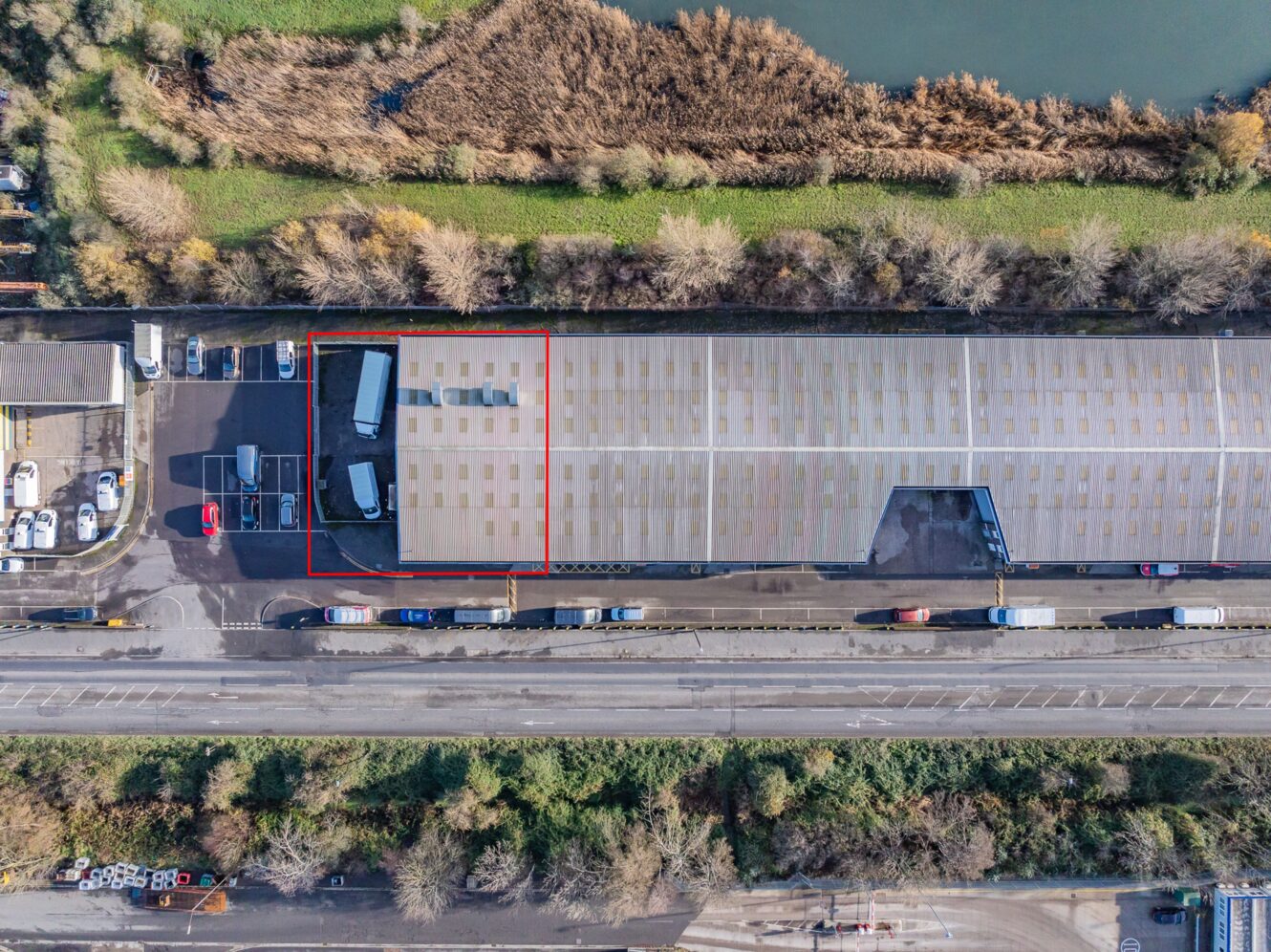Aerial view of a large warehouse with a red-outlined section, two lorries parked in front, and trees and roads surrounding the area.