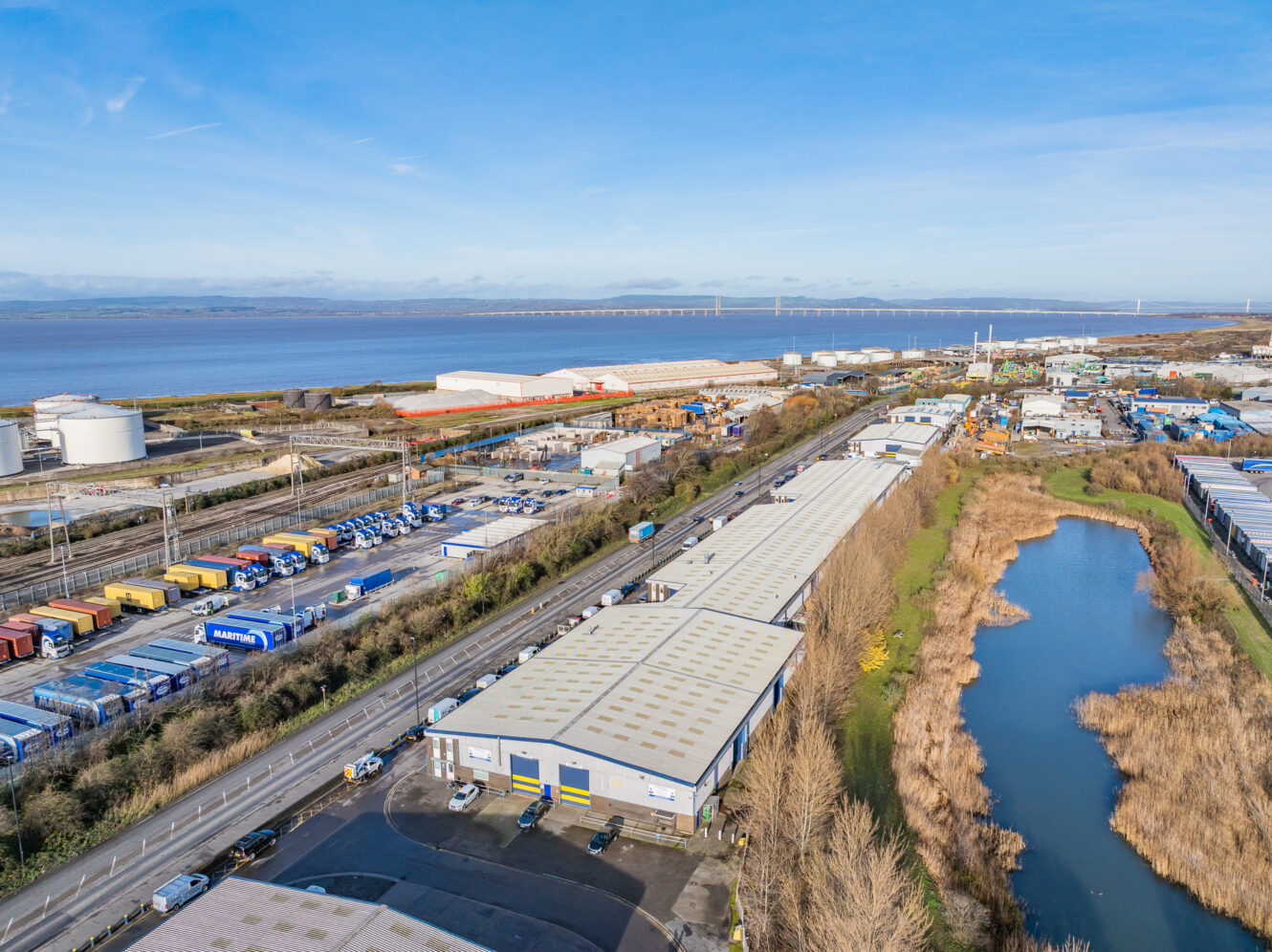 Aerial view of an industrial area with warehouses, parked lorries, a nearby waterway, and a bridge in the background under a blue sky.