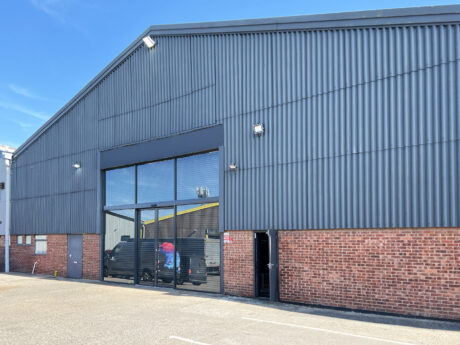 Industrial warehouse featuring corrugated metal cladding, expansive glass entrance doors, brickwork along the lower façade, and a partially open side door beneath a clear blue sky.
