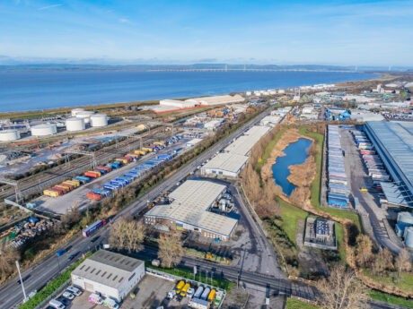Aerial view of an industrial area featuring warehouses, shipping containers, storage tanks, a pond, and railway tracks close to a coastline beneath a clear sky.