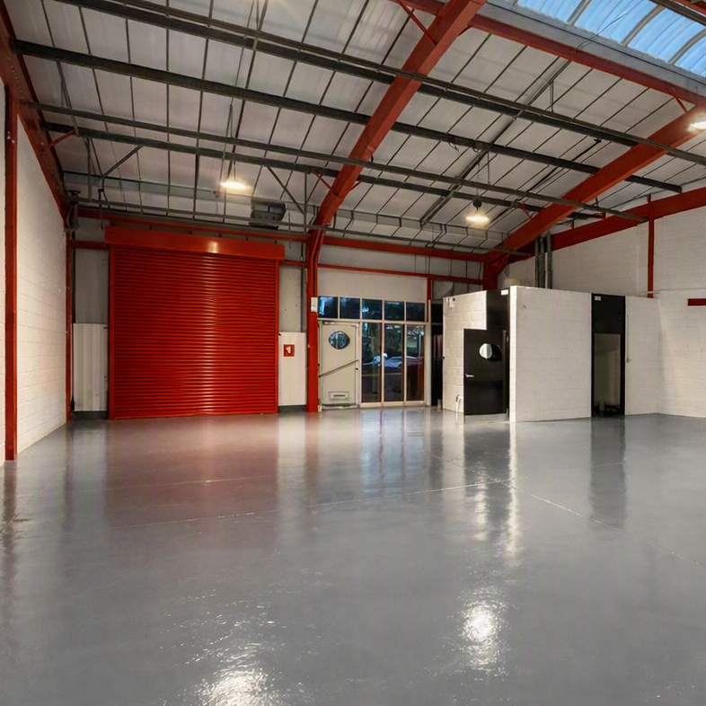 Interior of an empty industrial warehouse featuring a red roller shutter, white walls, a glossy grey floor, and a small white office area with windows.