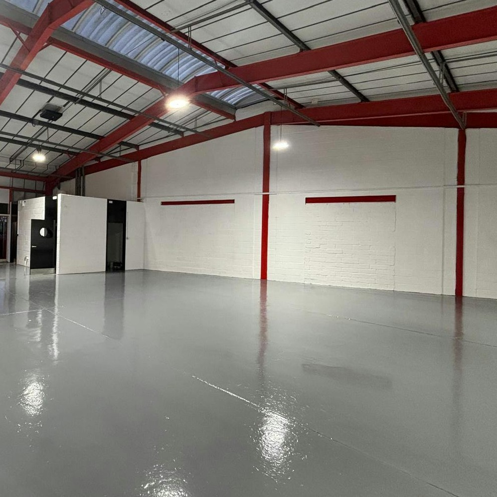 Large empty warehouse with a grey polished floor, white brick walls, red beams, and a metal ceiling. A small enclosed room is visible in the back corner. Bright overhead lighting fills the space.