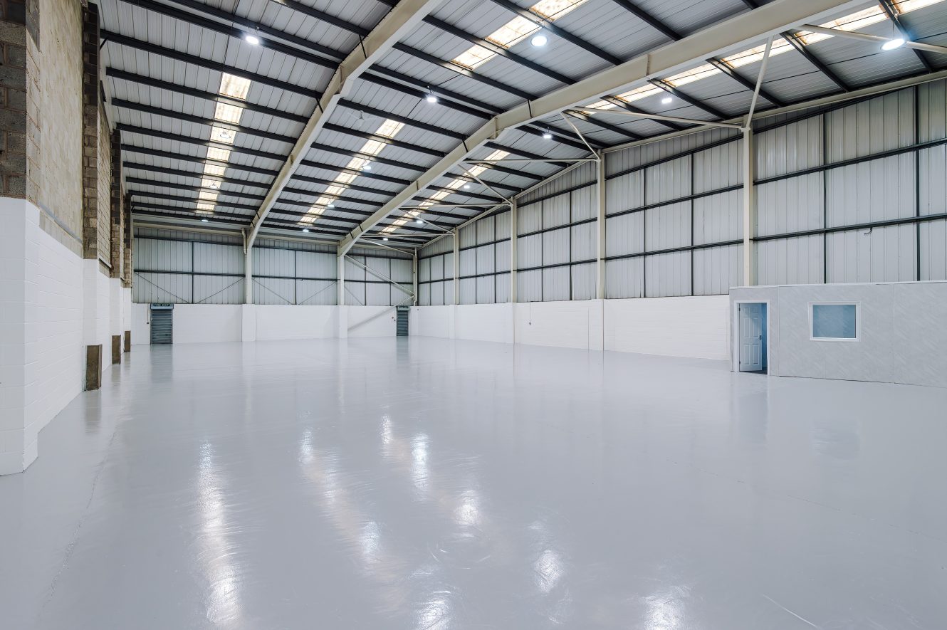 Large, empty industrial warehouse with polished concrete floors, high ceilings, exposed beams, white walls, and a small office section on the right side. Bright overhead lighting.
