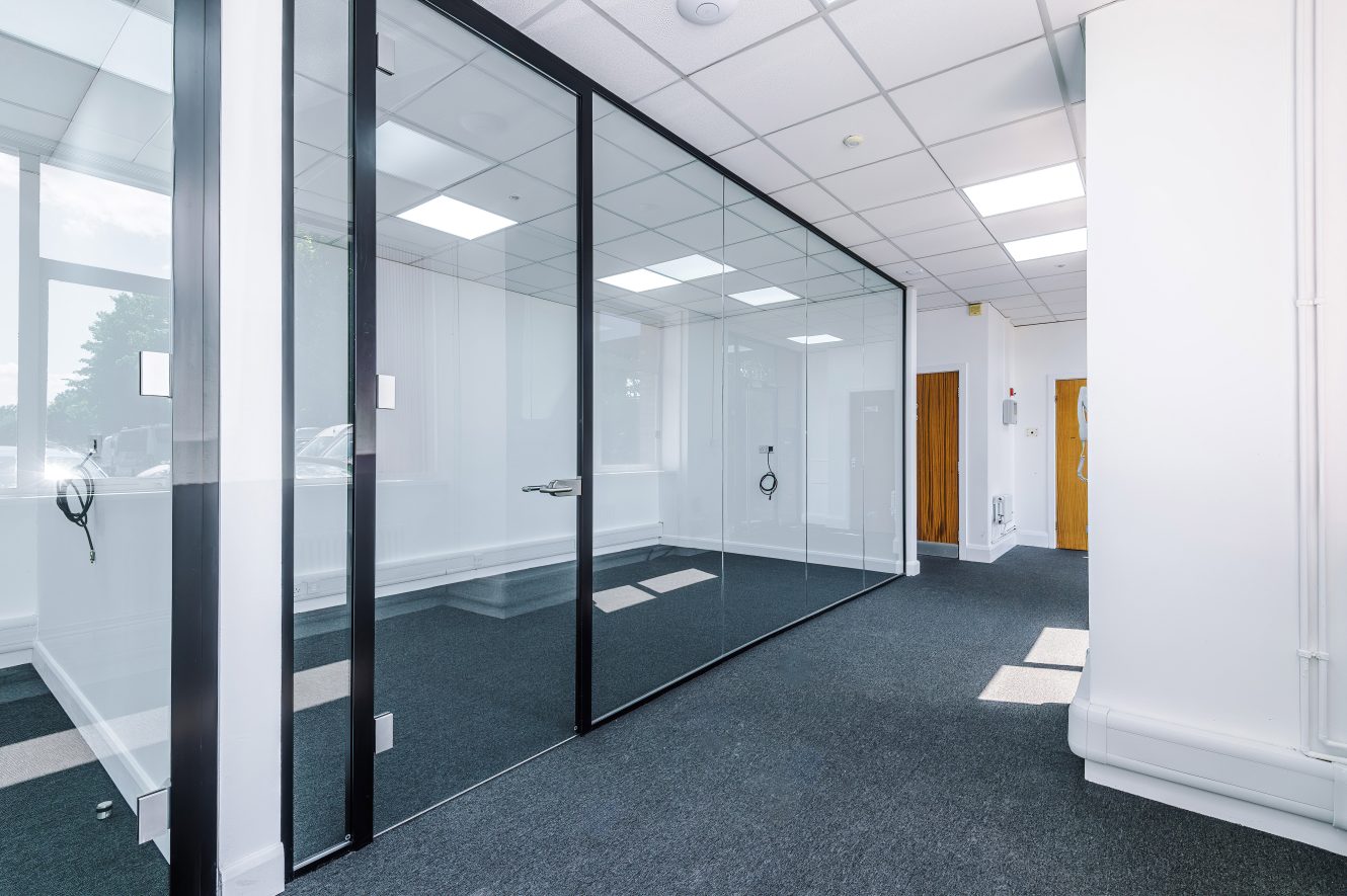 Modern empty office space featuring glass-walled meeting rooms, white walls, grey carpet, and ceiling lights.