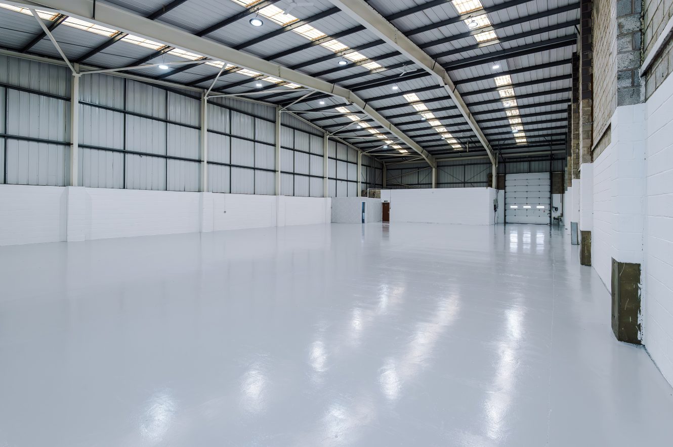 Large empty industrial warehouse with polished concrete floor, white walls, high ceiling, and metal beams, illuminated by skylights and overhead lights.