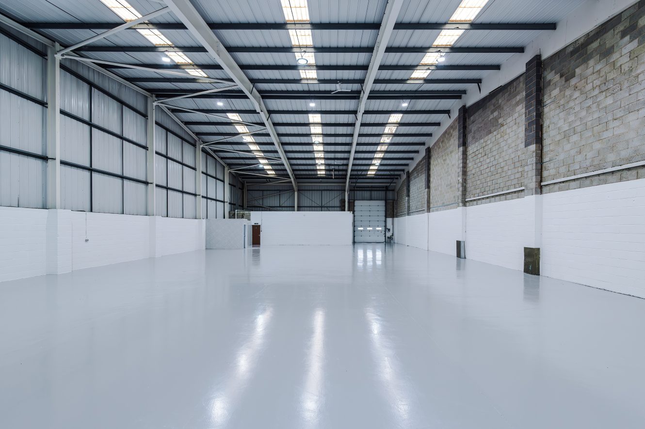 Empty industrial warehouse with high ceilings, white walls, exposed beams, and polished concrete floor, lit by rows of fluorescent lights.