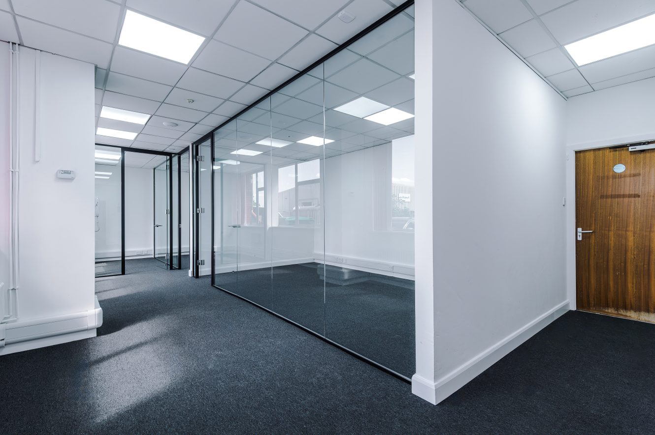 Modern office space with a glass-walled meeting room, grey carpet, white walls, and a wooden door; ceiling features fluorescent lights.