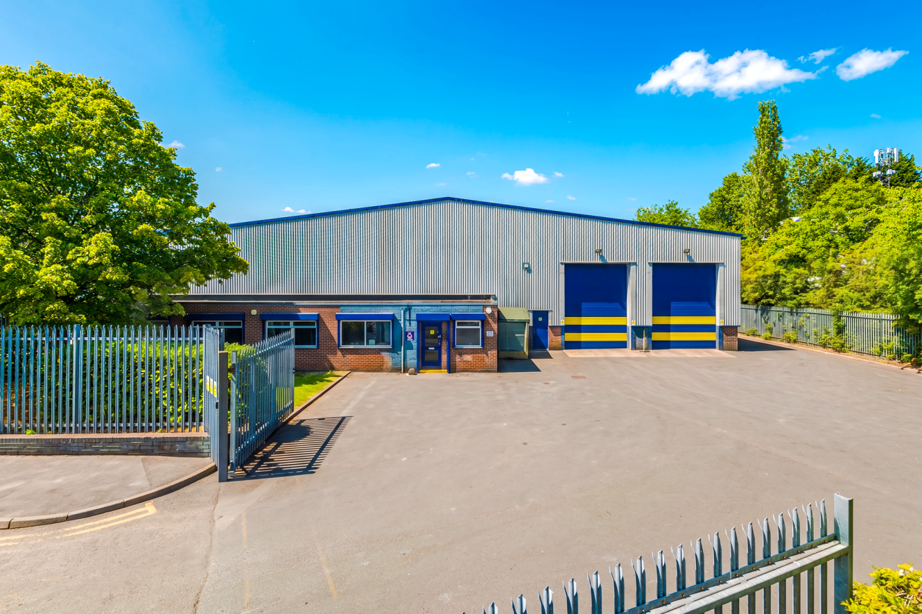 Industrial warehouse with two blue and yellow loading doors, a fenced gate, a large paved yard, and trees surrounding the building under a clear blue sky.
