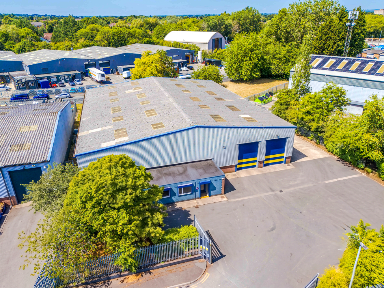 A large industrial warehouse with a gated entrance, surrounded by trees and other industrial buildings under a clear blue sky.