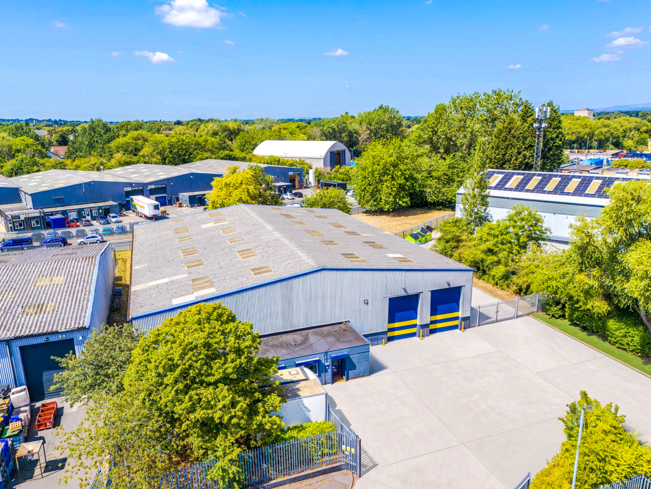 Aerial view of an industrial warehouse with a grey roof, large car park, and surrounding trees on a clear day.