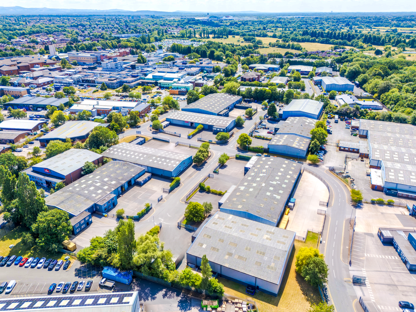 Aerial view of an industrial estate with warehouses, office buildings, roads, parked vehicles, and green areas on a sunny day.