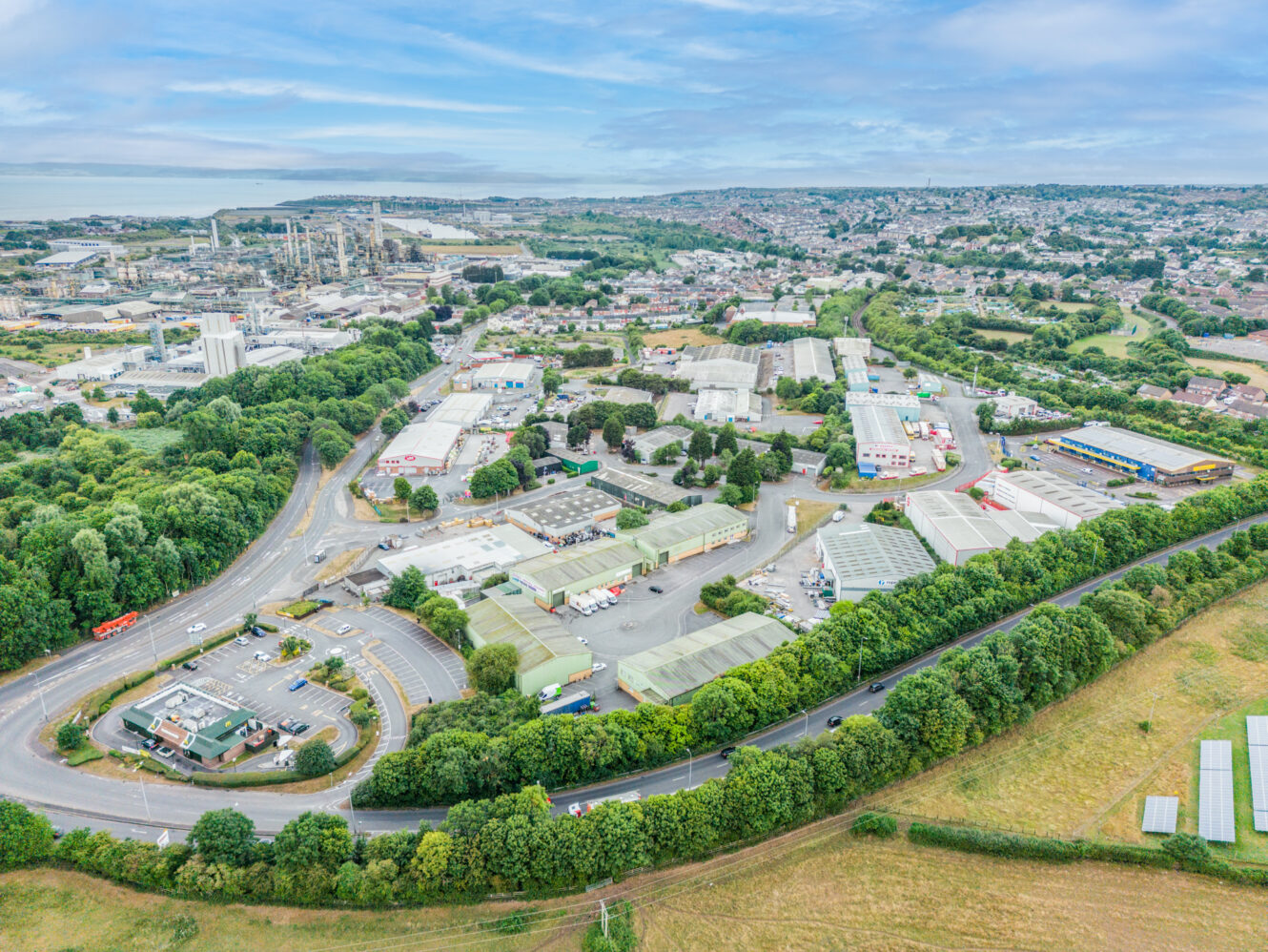 Aerial view of an industrial estate featuring warehouses and roads bordered by green trees and fields, with a residential area and coastline visible in the background beneath a partly cloudy sky.