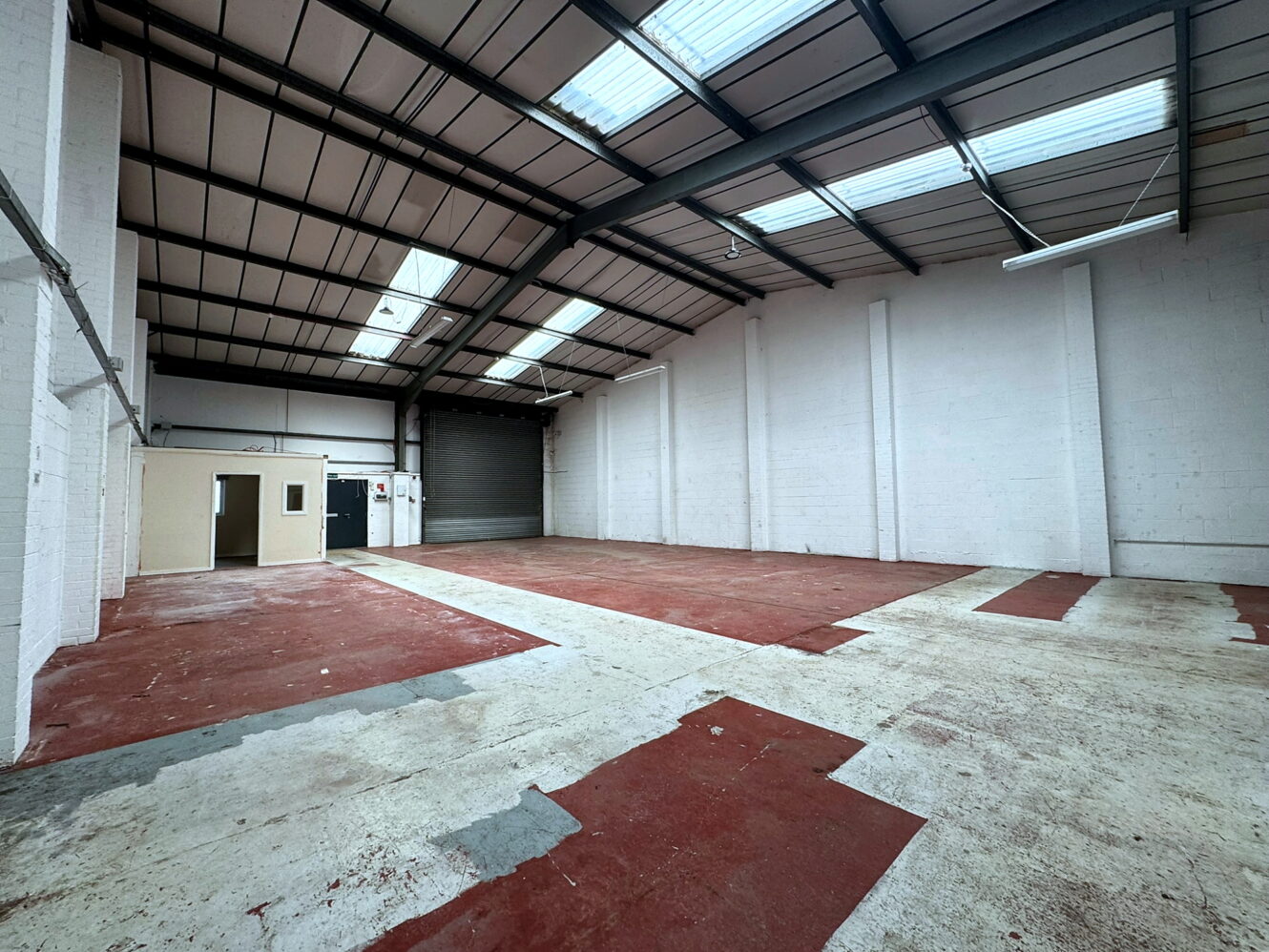 Empty industrial warehouse interior with high ceiling, skylights, white brick walls, red and white painted concrete floor, and a partially open roller shutter at the back.