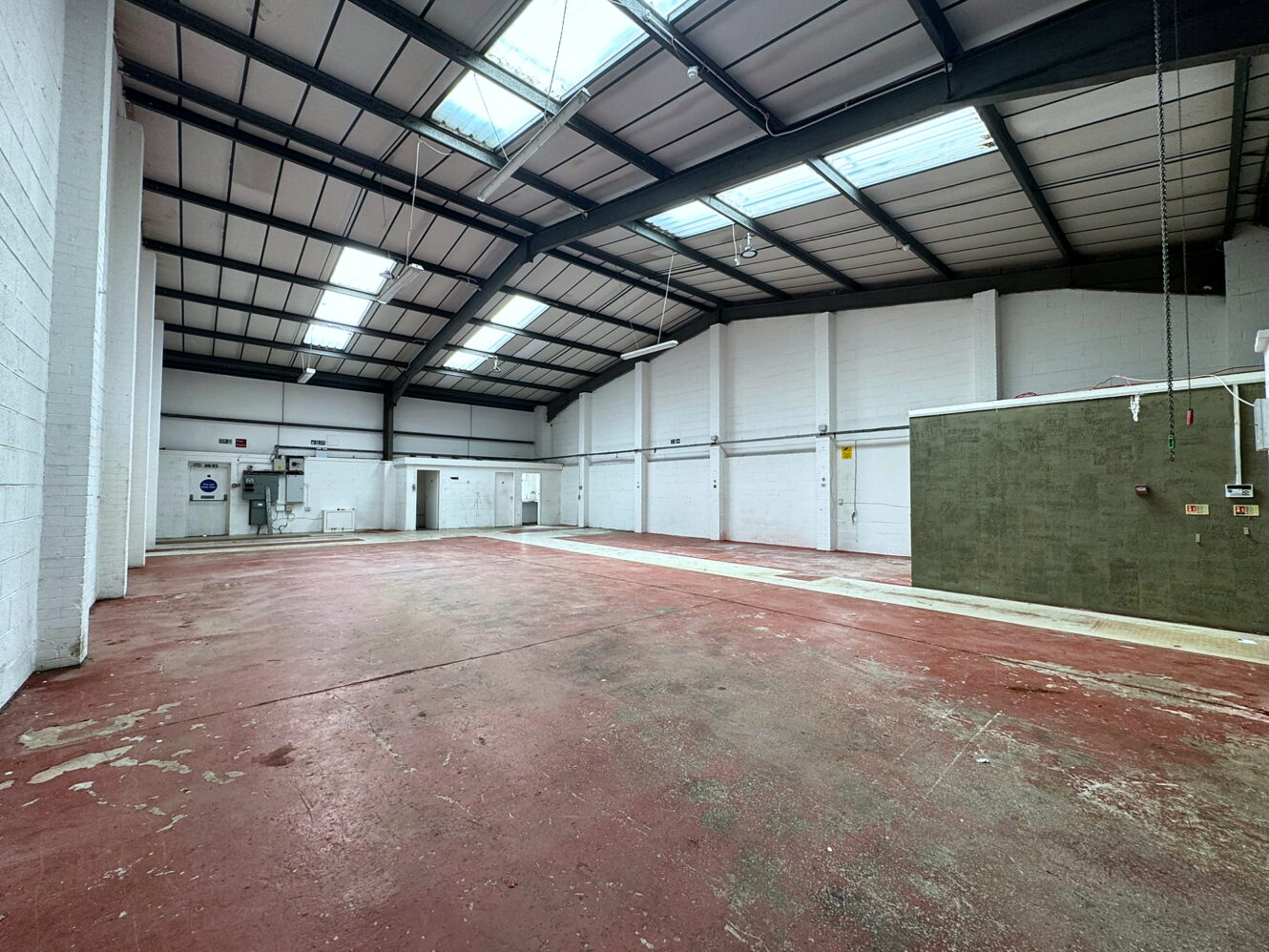 Large, empty industrial warehouse with a red concrete floor, white brick walls, high ceiling, skylights, and utility boxes along the walls.
