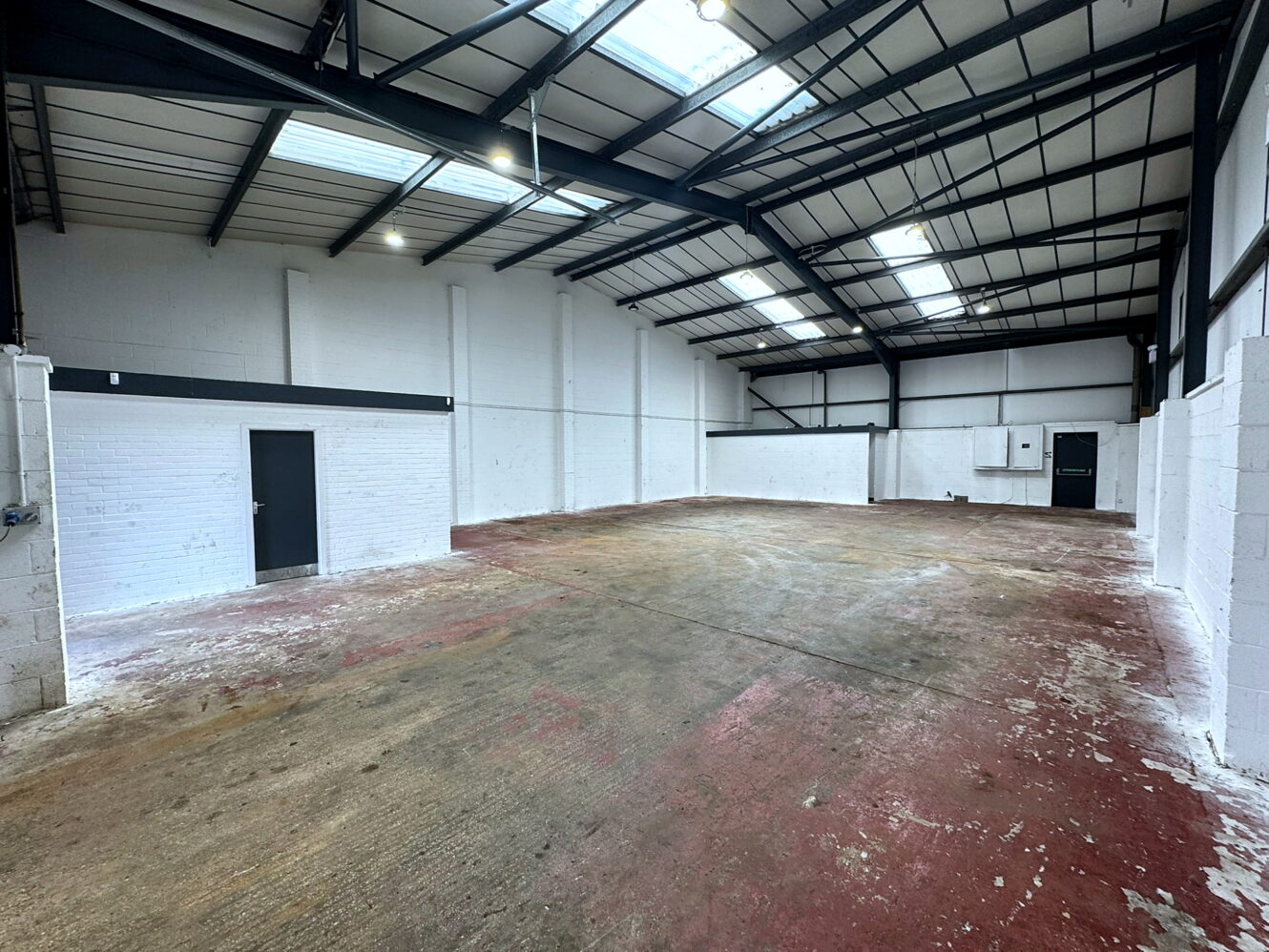Empty industrial warehouse with white brick walls, exposed steel beams, skylights, and two black doors; the floor appears worn and stained.