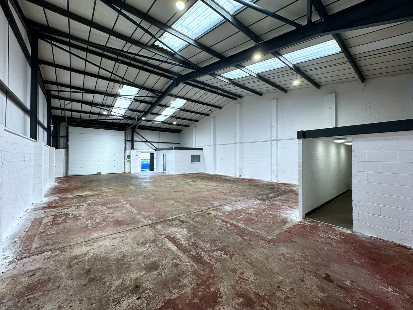Large empty warehouse with high ceilings, skylights, white walls, concrete floor, and a small office area visible to the right.