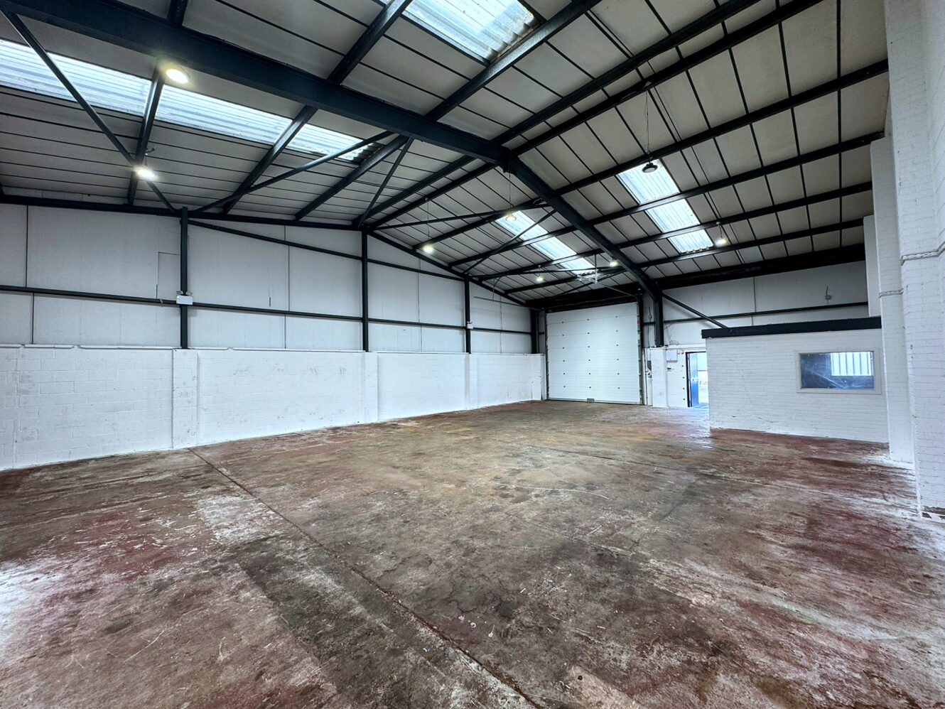 Empty industrial warehouse with high ceilings, exposed beams, white walls, large garage door, and concrete floor, lit by skylights and ceiling lights.