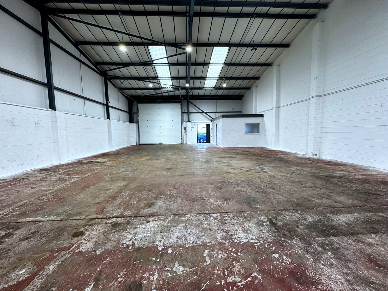 Empty industrial warehouse with high ceilings, exposed beams, white walls, concrete floor, a small office window, and a large garage door at the far end.