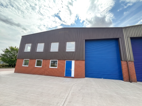 A modern industrial building with red brick and dark cladding, featuring a blue door and a large blue roller shutter, under a partly cloudy sky.