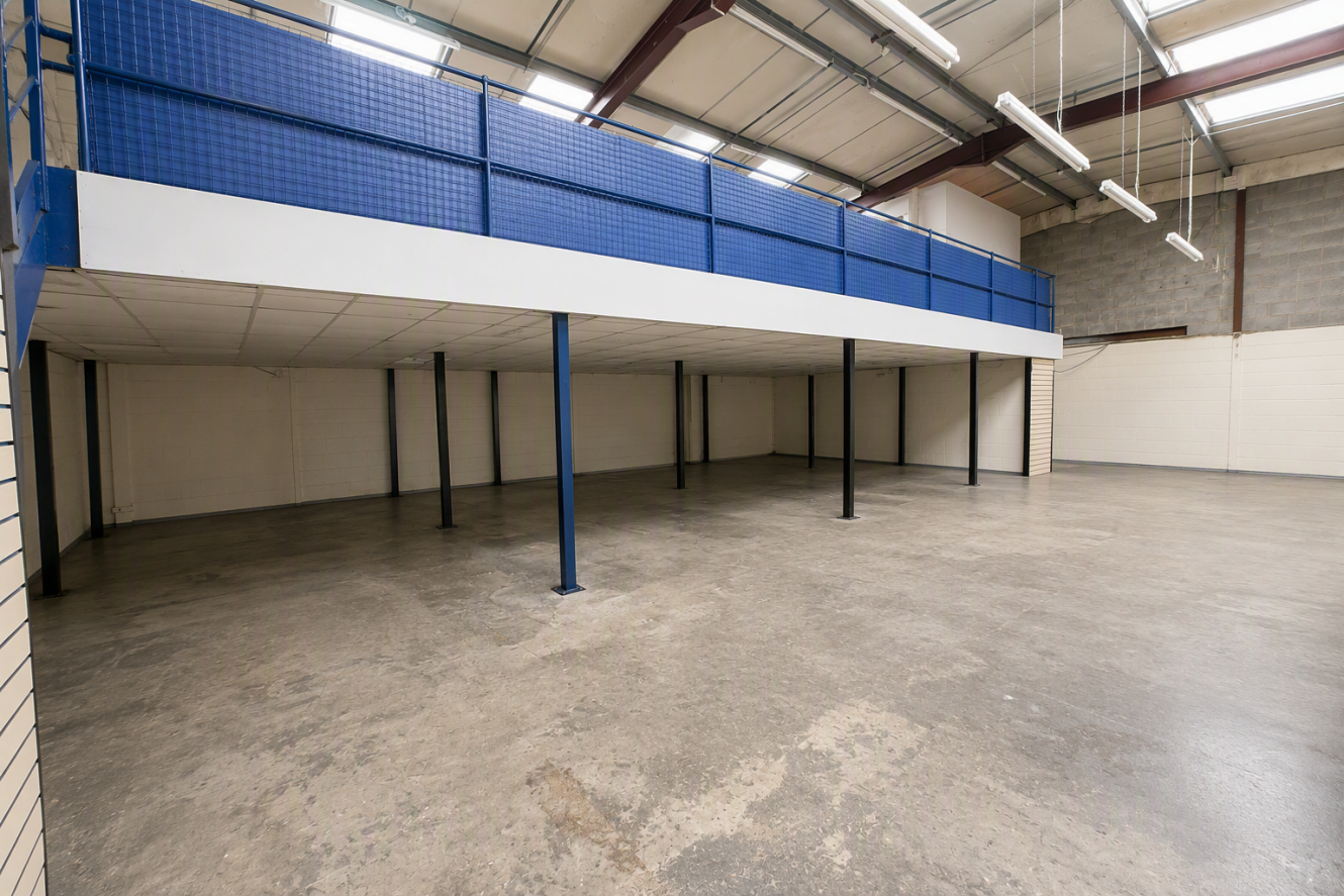 Empty warehouse interior with a concrete floor, white walls, exposed ceiling beams, and a blue metal mezzanine supported by black pillars, all brightly lit by rows of hanging fluorescent lights.