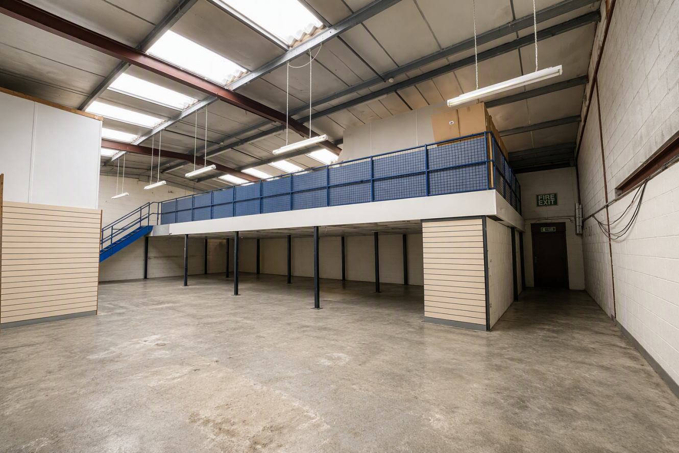 Spacious empty warehouse interior with polished concrete floor, mezzanine level, blue stairs, and fluorescent lighting. Walls are white with beige panelling.
