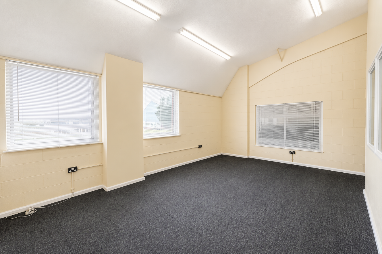 Empty office room with beige walls, three large windows covered by blinds, and grey carpet flooring illuminated by bright ceiling lights.