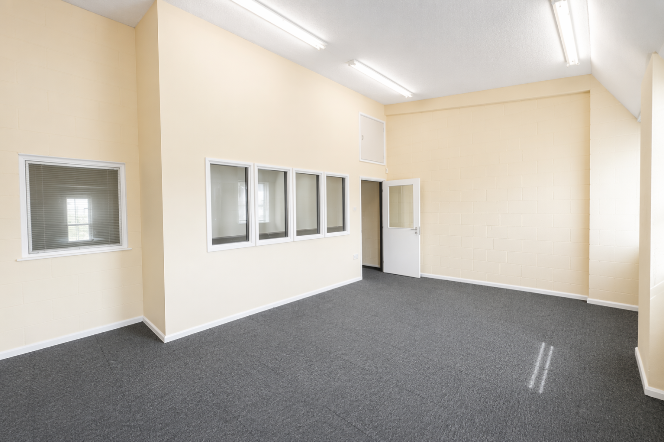 Empty office room with beige walls, grey carpet, several interior windows, a closed white door, and fluorescent lighting on the ceiling.