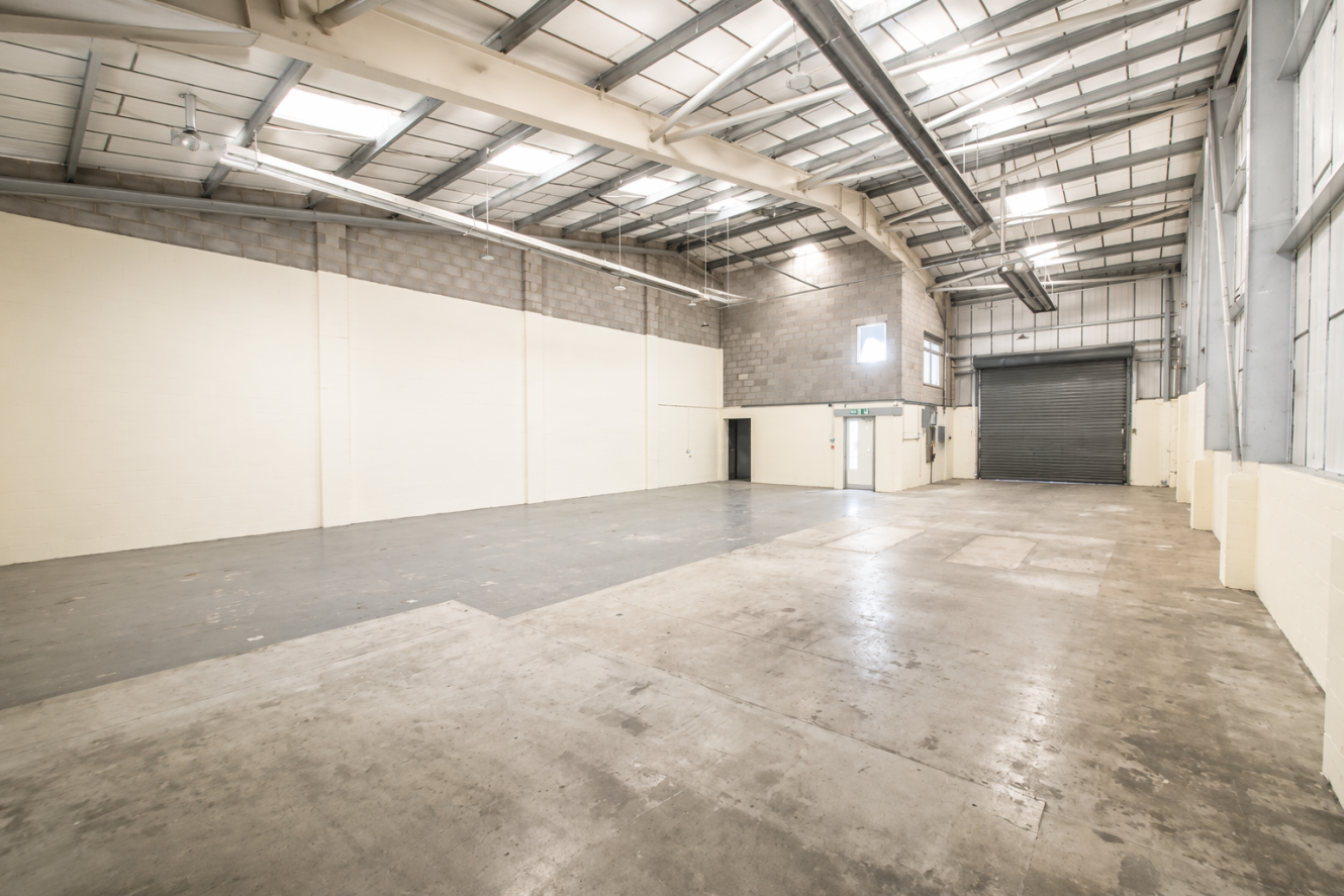 Spacious empty warehouse with concrete floors, high ceilings, beige walls, overhead lights, and a large roller shutter at the far end.