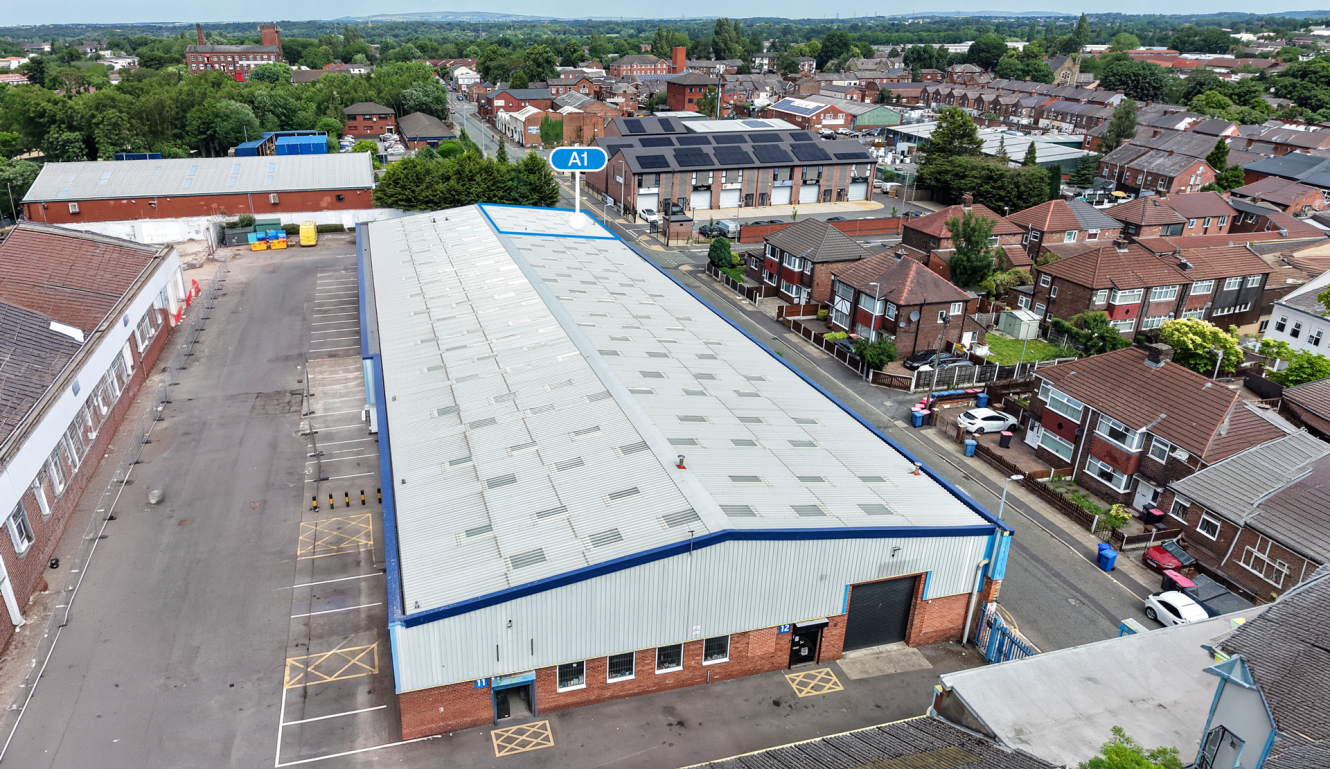 A large industrial warehouse with a white roof is surrounded by residential houses and other buildings in a suburban area.