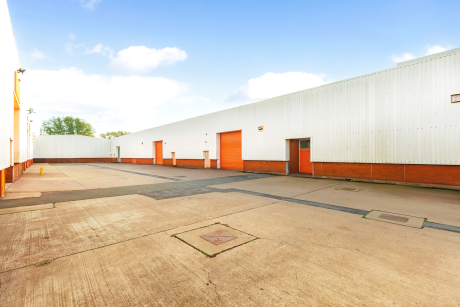 Empty industrial warehouse yard with white and orange buildings, multiple garage doors, and a clear sky overhead.
