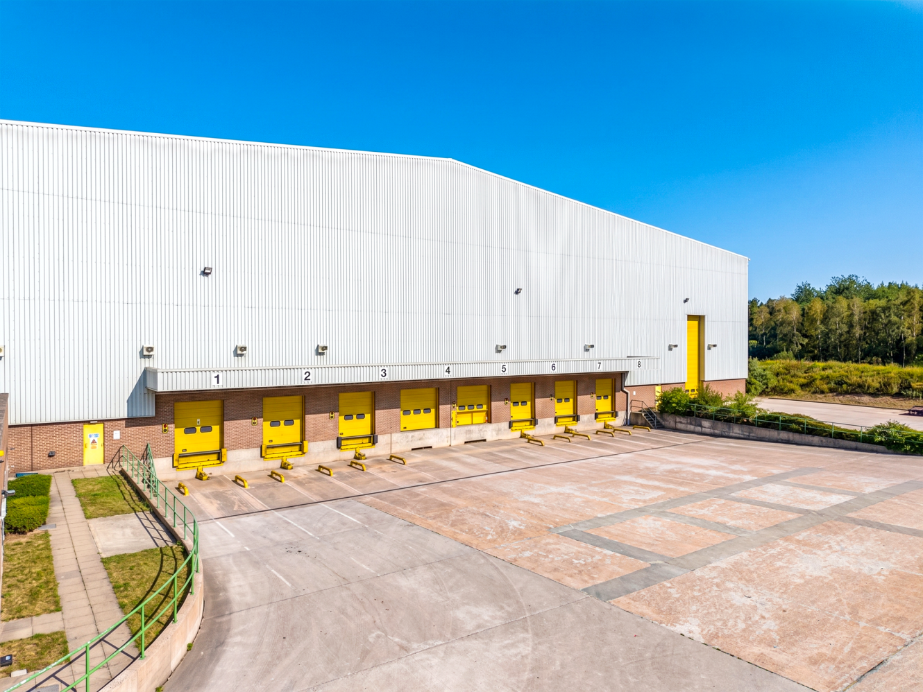 Large white industrial warehouse with six yellow loading bay doors, a concrete loading area, and a clear blue sky. Trees and greenery are visible in the background.