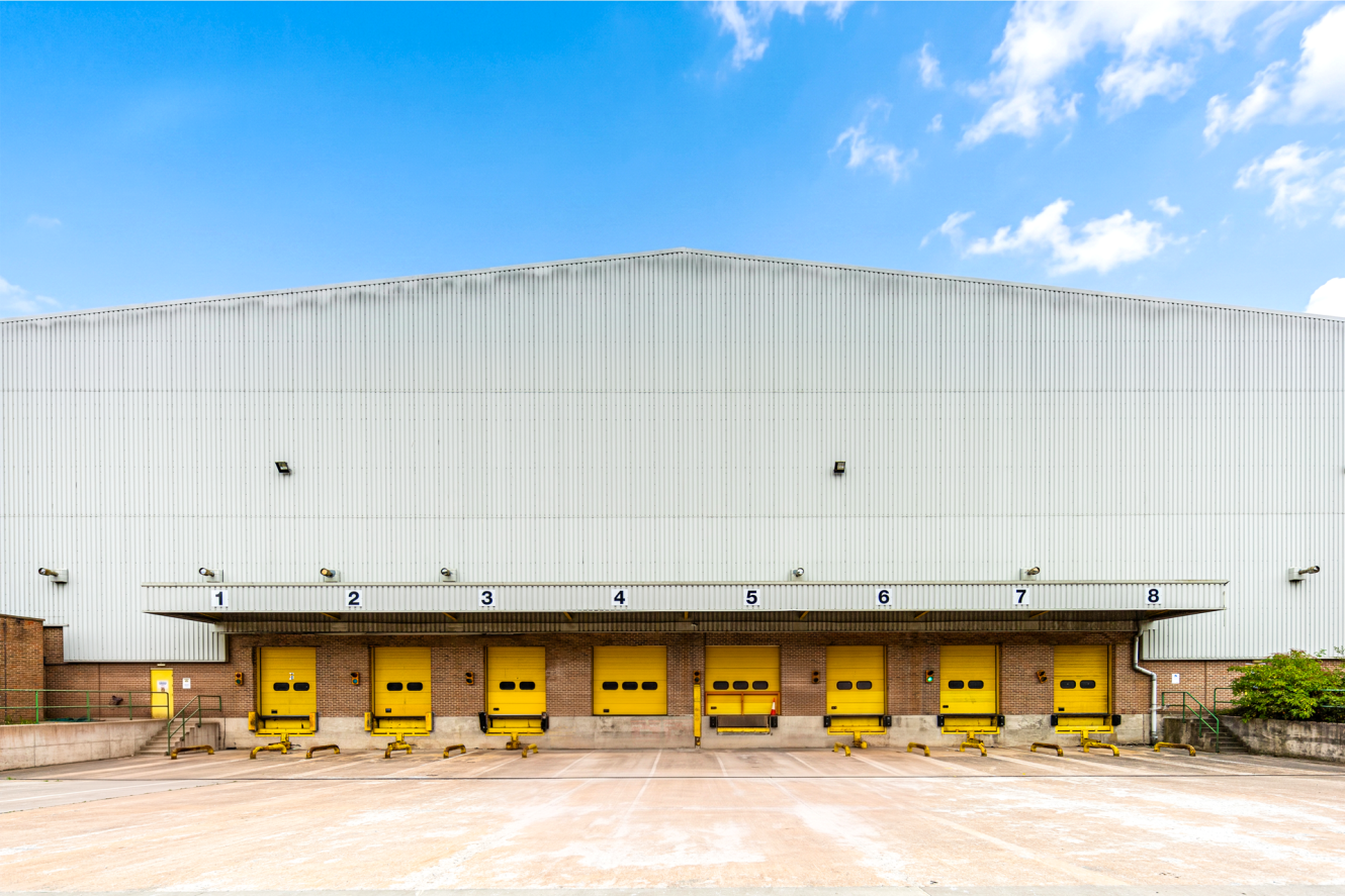 A warehouse exterior with eight numbered yellow loading bay doors under a corrugated metal roof, seen on a clear day with blue sky and clouds.