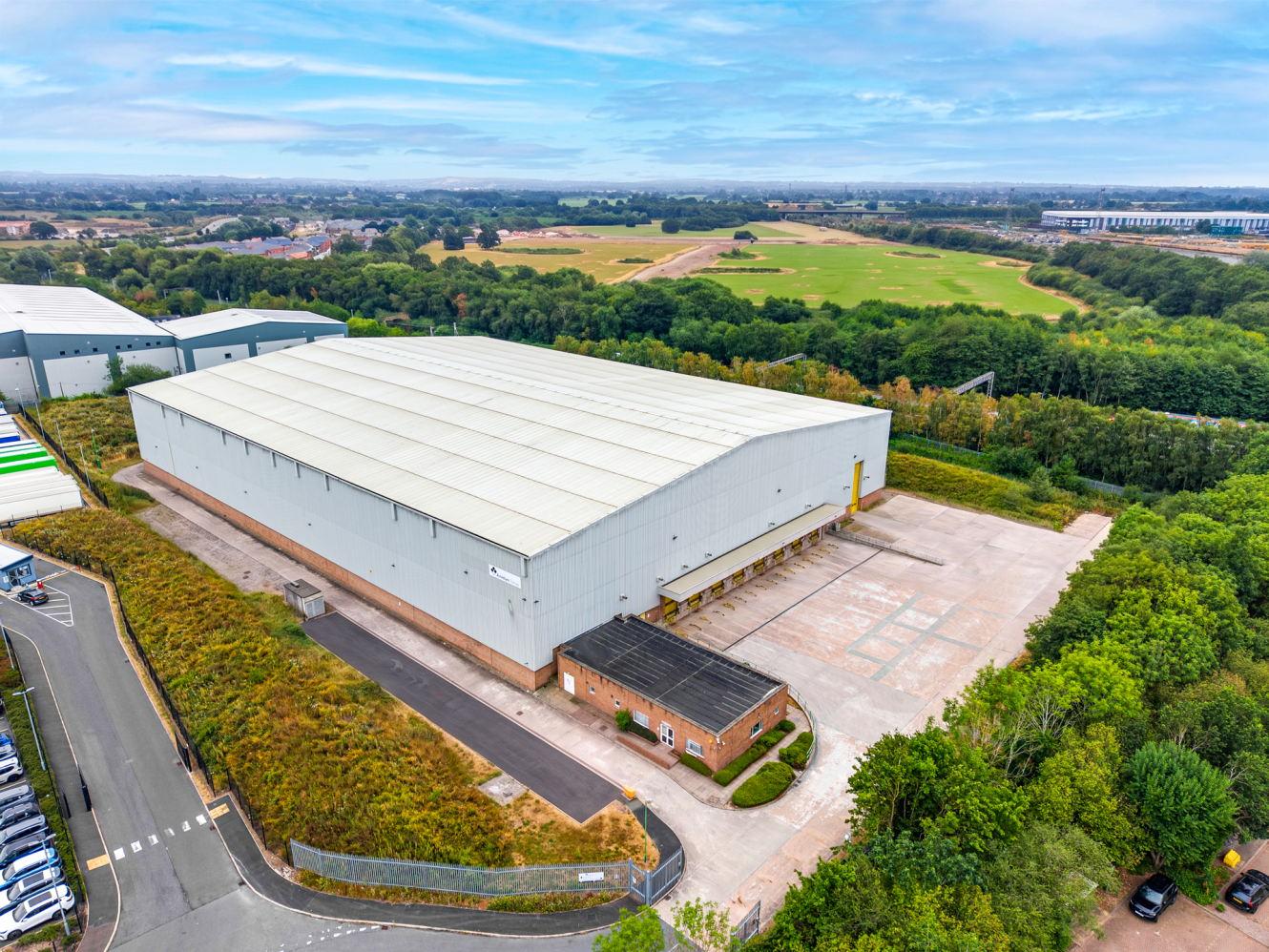 A large warehouse with a white roof is surrounded by trees and open fields, with a small brick building and a car park in front.