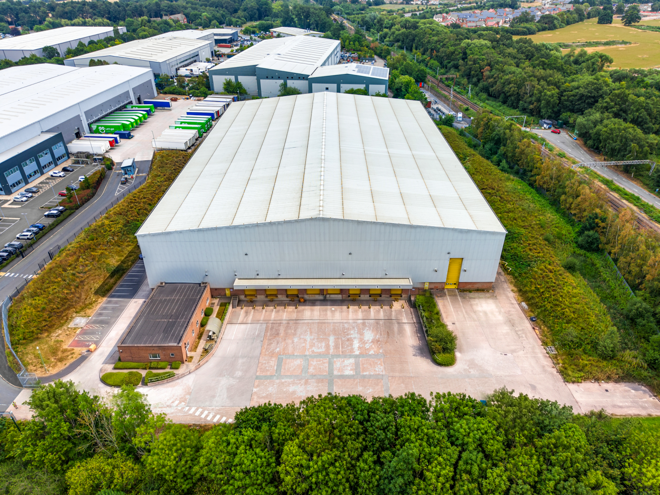 Aerial view of a large warehouse with a white roof, loading bays, parking area, and surrounding greenery in an industrial estate.