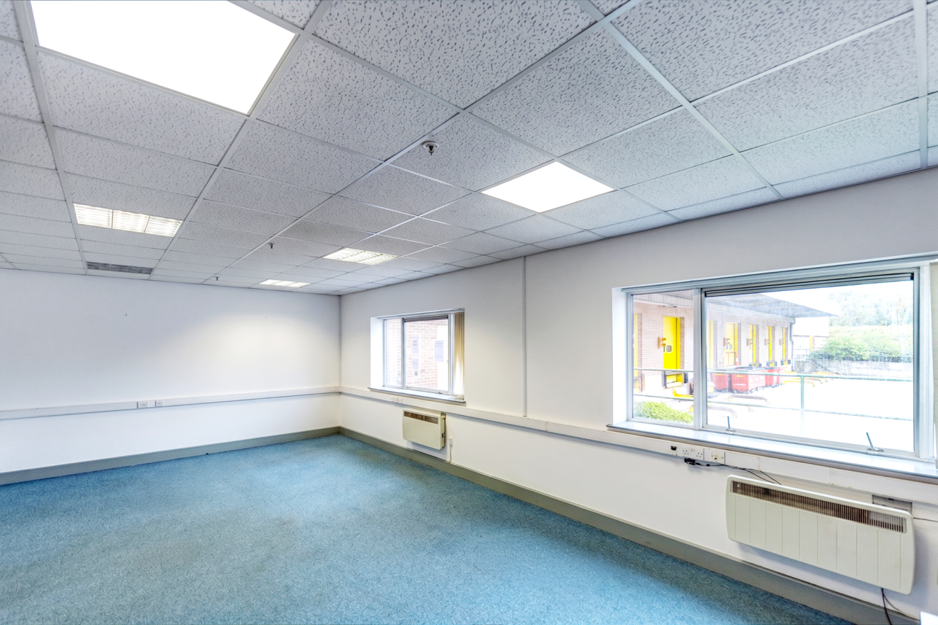 Empty office room with blue carpet, white walls, ceiling lights, two windows, and visible wall heaters.