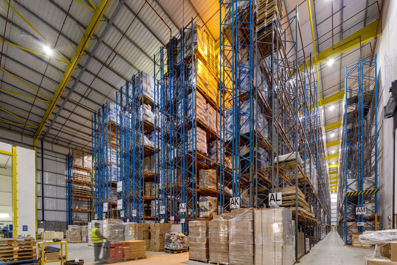 Large warehouse with tall blue racks filled with pallets of goods. Someone in a safety vest operates a pallet truck. The space is well-lit and organised.