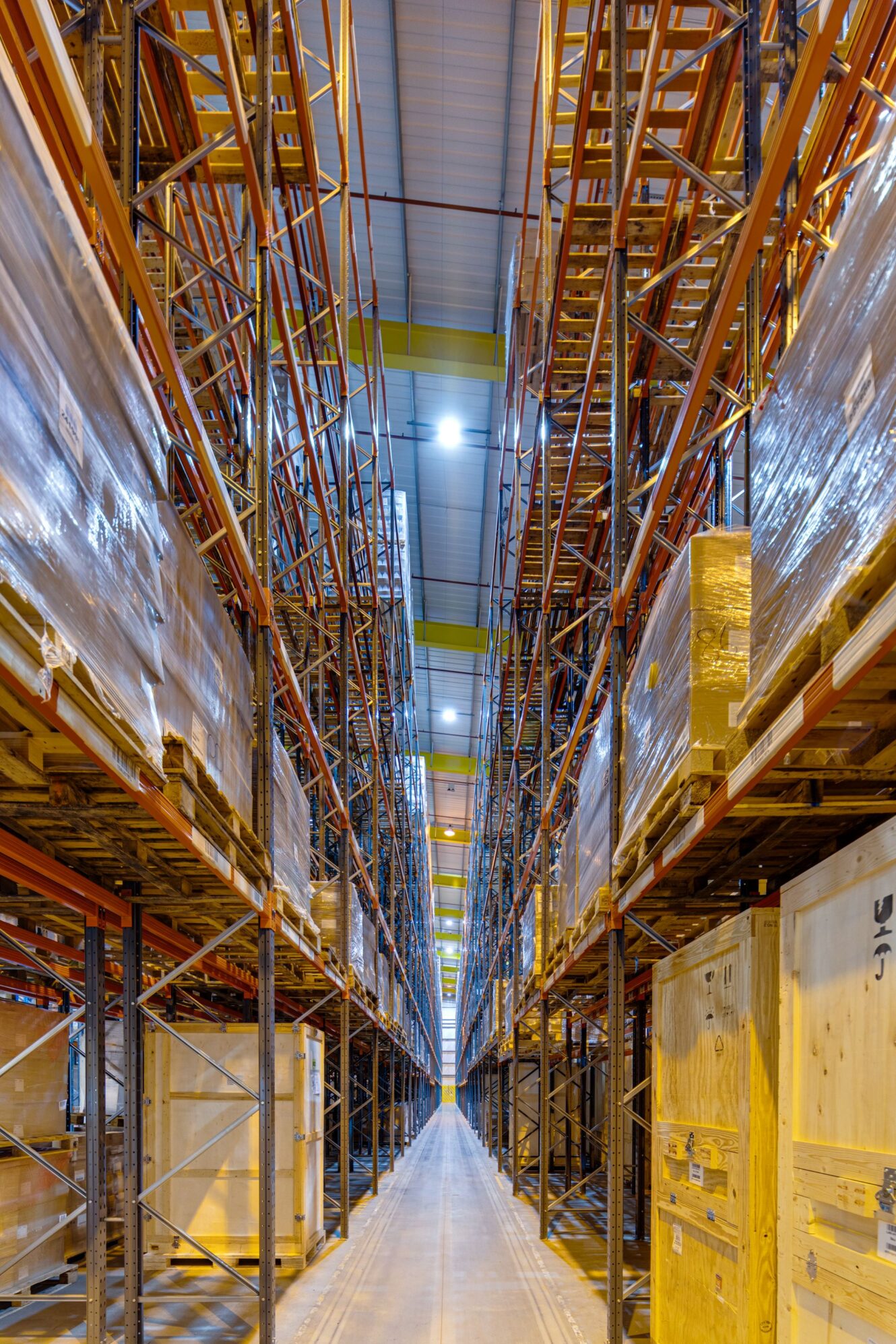 Wide-angle view of tall, organised warehouse shelves filled with stacked pallets, wooden crates, and various boxed materials under bright overhead lighting.